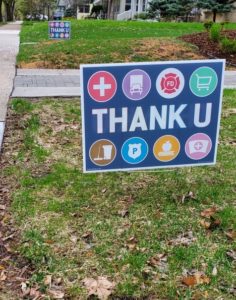 A "Thank U" sign with colorful symbols for essential workers sits is stuck in the grass by a sidewalk.